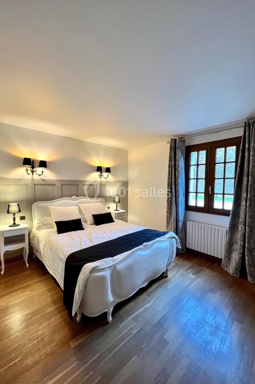 Chambre lumineuse avec lit double, tables de chevet, lampes, parquet en bois et fenêtre avec rideaux gris.