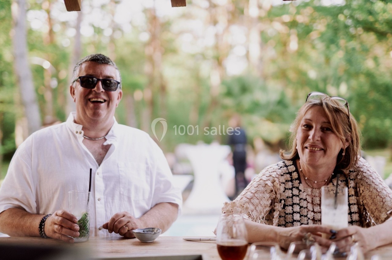 Un homme et une femme sourient assis à une table en extérieur, avec un fond de végétation et une piscine floue.