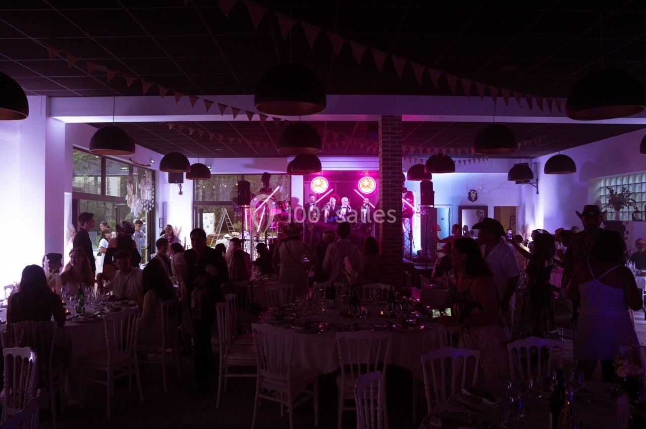 Salle de réception décorée avec des guirlandes, des invités assis et un groupe jouant sur une scène éclairée en rose.
