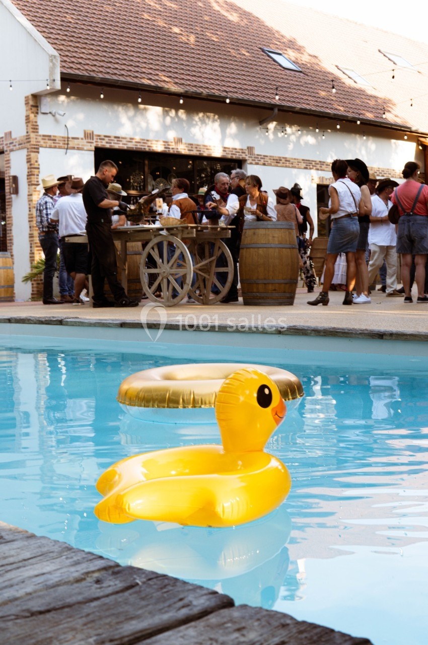 Canard gonflable jaune flottant dans une piscine, avec des personnes rassemblées près d'un bar en arrière-plan.