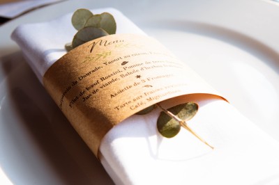 Serviette blanche pliée avec un menu enroulé et une branche d'eucalyptus, posée sur une assiette blanche.