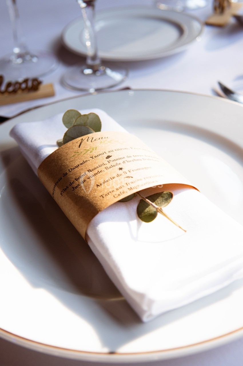 Serviette blanche pliée avec un menu enroulé et une branche d'eucalyptus, posée sur une assiette blanche.