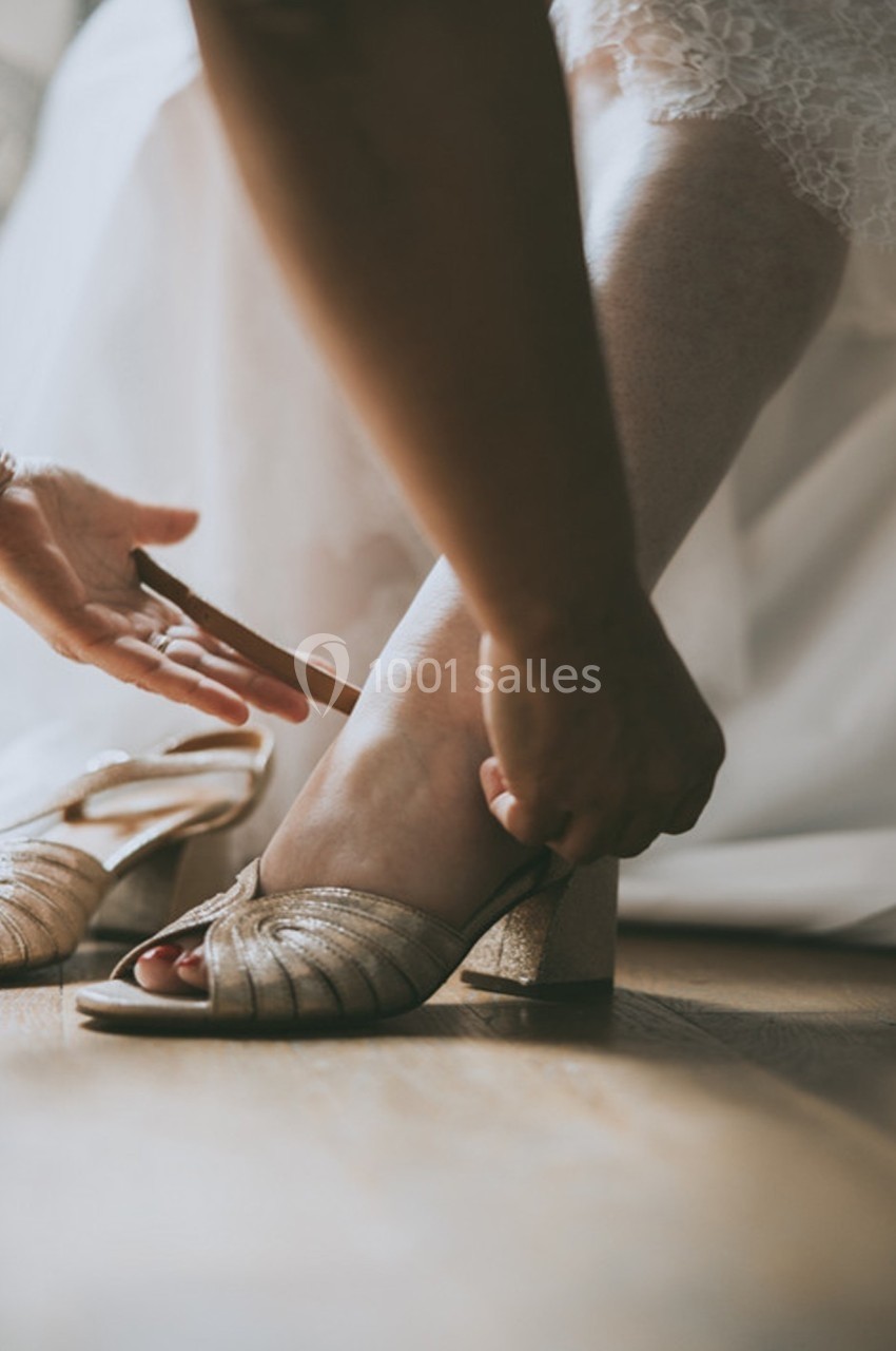 Une personne ajuste une chaussure dorée à talon sur le pied d'une femme en robe blanche.