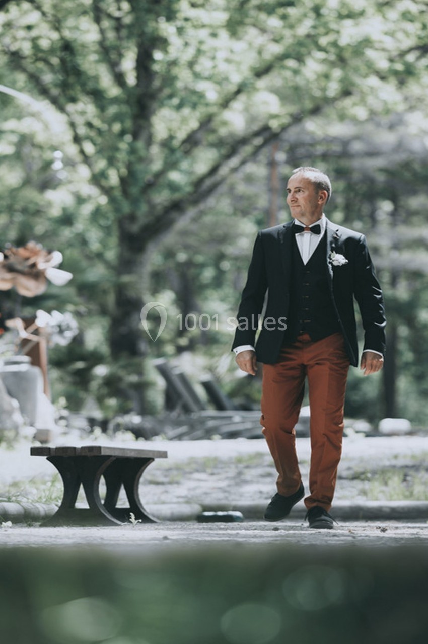 Un homme en costume marche dans un jardin boisé, près d'un banc, sous une lumière naturelle.