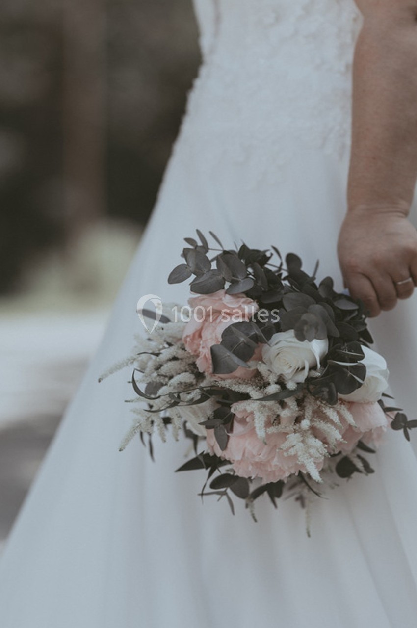 Une mariée en robe blanche tient un bouquet de fleurs roses et blanches avec des feuillages verts.