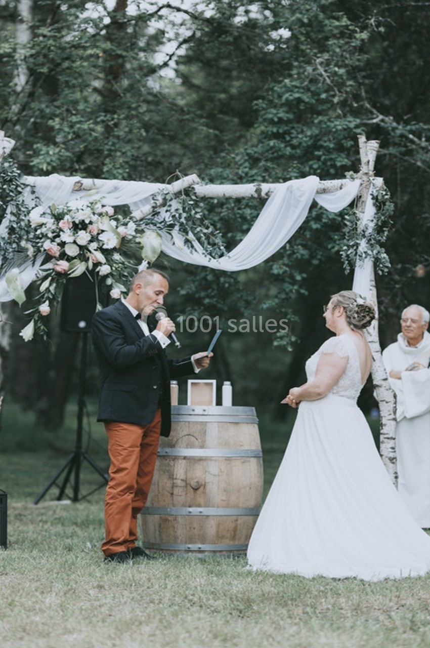 Un homme lit un texte lors d'une cérémonie de mariage en plein air, sous une arche décorée de fleurs et de voilages.