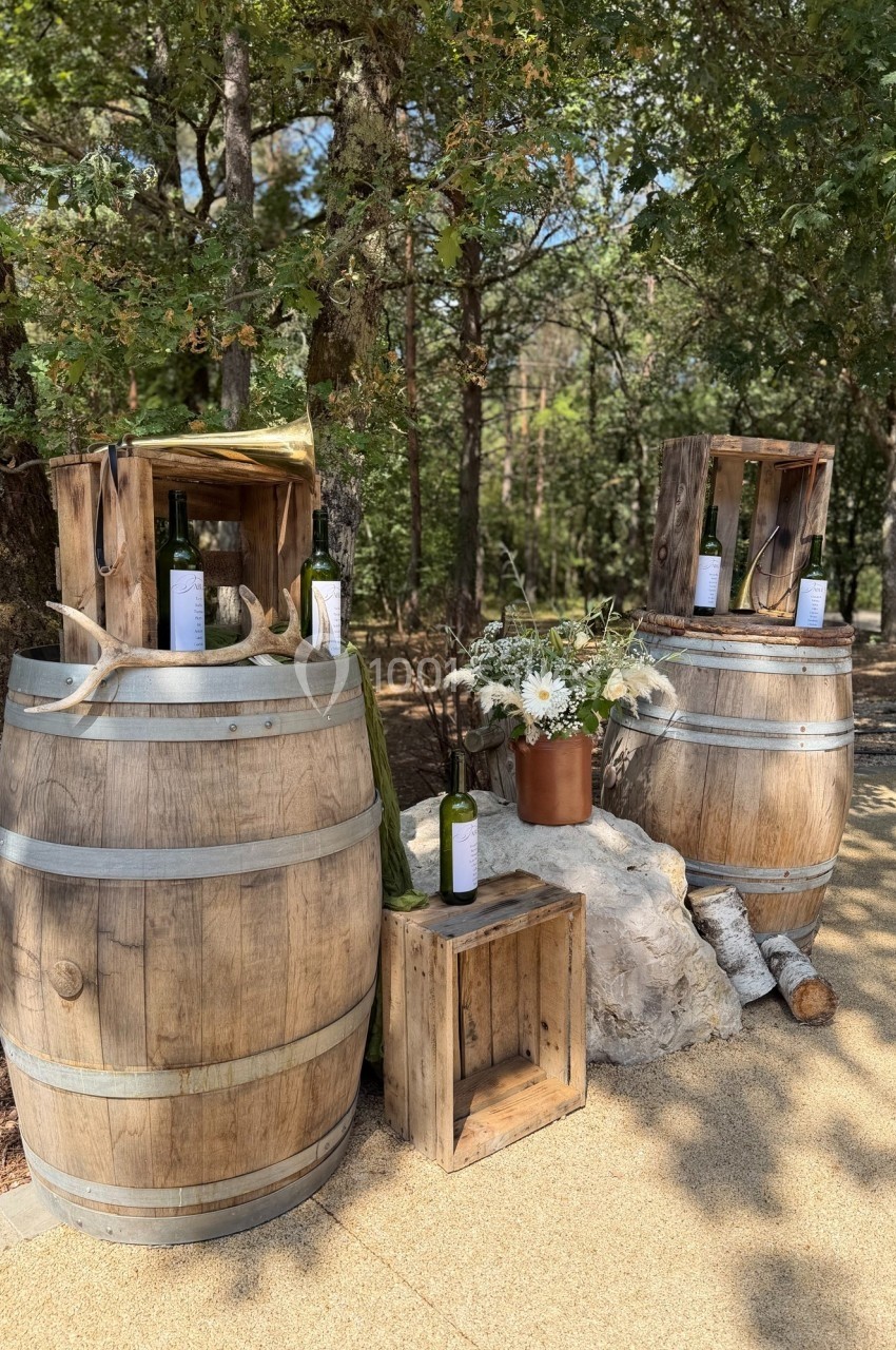 Deux tonneaux en bois décorés avec des bouteilles de vin, des fleurs et des caisses, dans un cadre extérieur boisé.