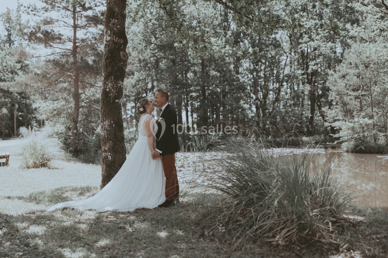 Un couple en tenue de mariage se tient sous un arbre près d'un étang dans un cadre naturel boisé.