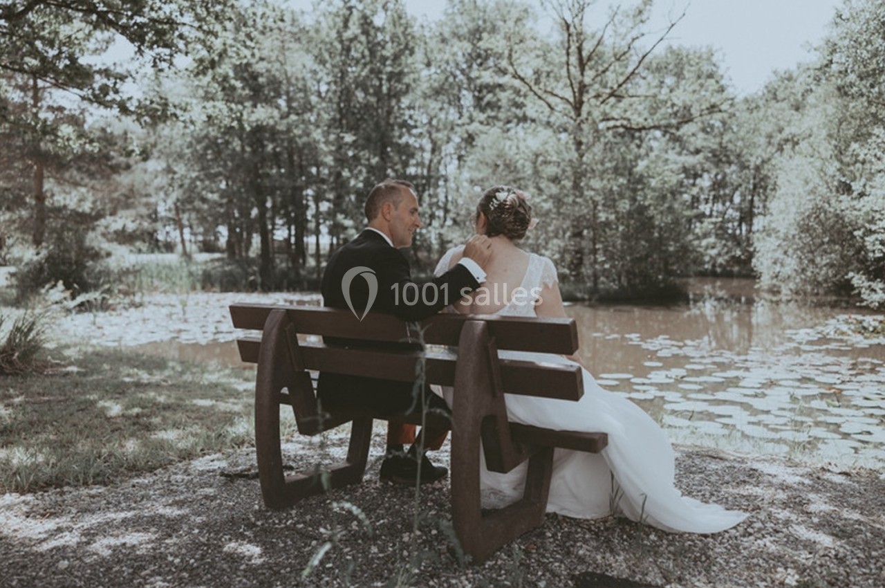 Un couple assis sur un banc en bois près d'un étang entouré d'arbres, échangeant un moment complice.