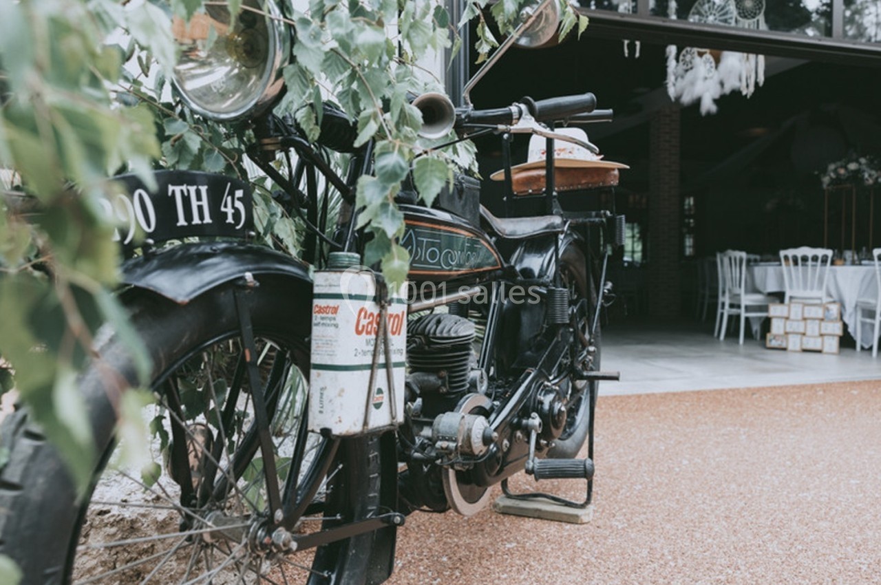 Ancienne moto stationnée devant une entrée vitrée, entourée de végétation et de mobilier intérieur visible.