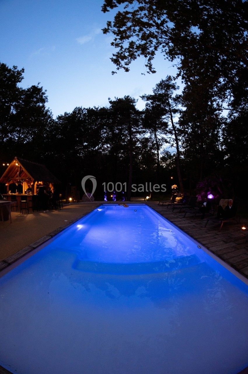 Piscine éclairée en soirée entourée d'arbres et d'un espace aménagé avec des lumières tamisées.