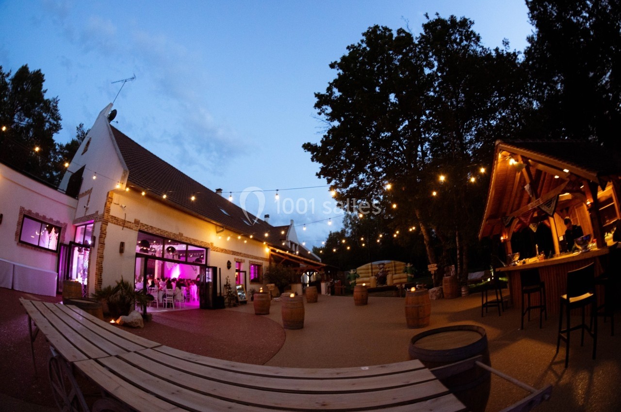 Cour extérieure éclairée avec guirlandes lumineuses, terrasse en bois, bar en plein air et bâtiment illuminé au crépuscule.