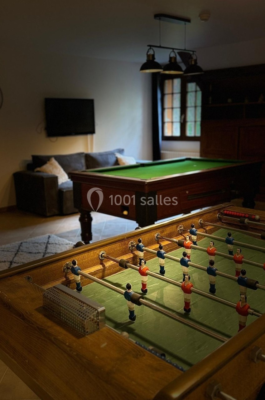 Baby-foot au premier plan avec un billard en arrière-plan dans une pièce aménagée avec canapé et télévision.