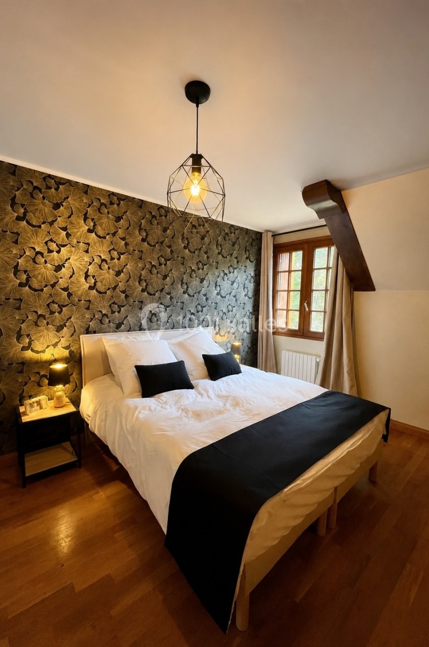 Chambre lumineuse avec lit double, linge blanc, coussins noirs, mur à motif floral sombre et fenêtre en bois.