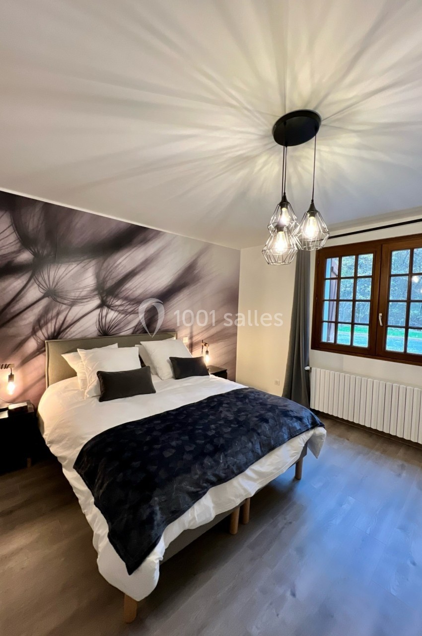 Chambre moderne avec lit double, suspension lumineuse design, mur décoratif et fenêtre donnant sur un extérieur verdoyant.