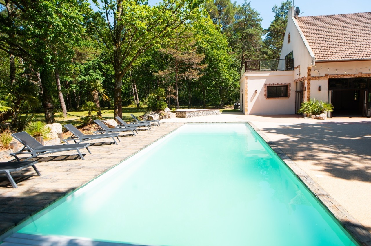 Piscine extérieure entourée de chaises longues, située dans un jardin arboré près d'une maison blanche.