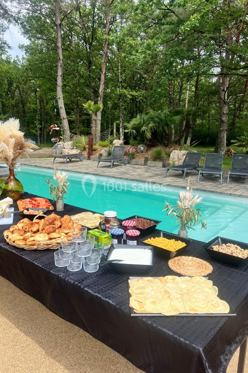 Table dressée avec des plats variés et des boissons, au bord d'une piscine entourée de végétation et de transats.