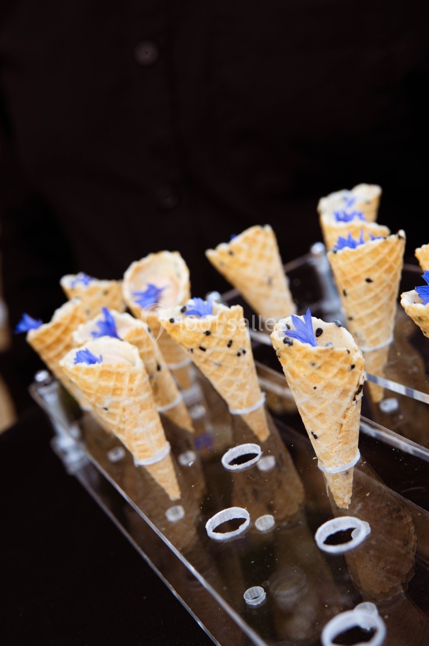 Cônes en biscuit garnis de crème et décorés de fleurs bleues, présentés sur un support transparent.
