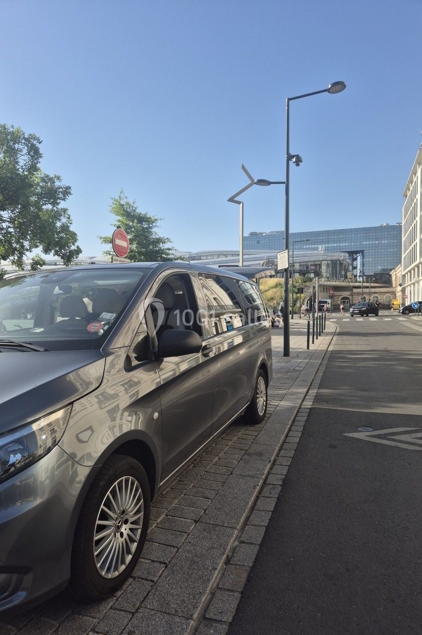 Un van gris stationné le long d'un trottoir dans une rue urbaine avec des bâtiments modernes en arrière-plan.