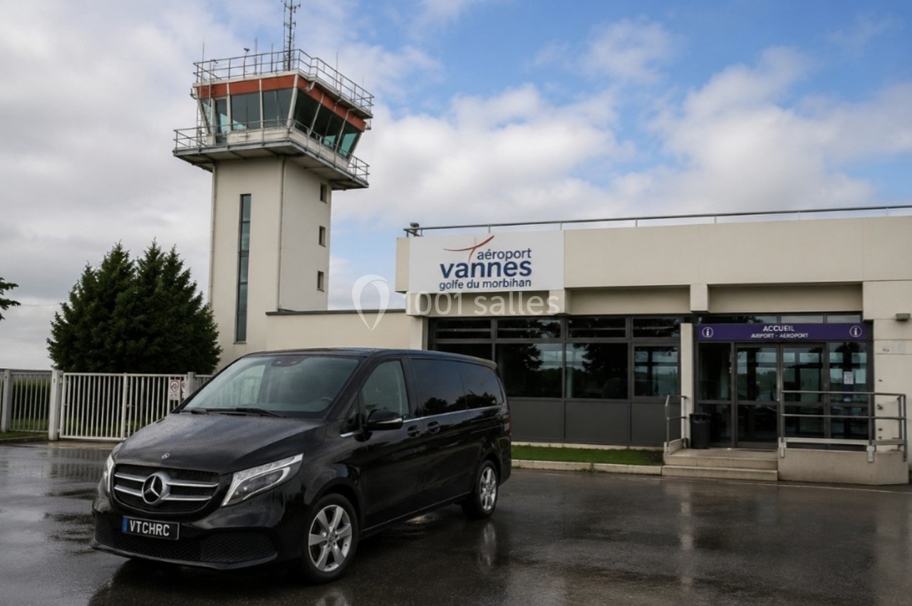 Minibus noir stationné devant l'entrée de l'aéroport de Vannes avec une tour de contrôle en arrière-plan.