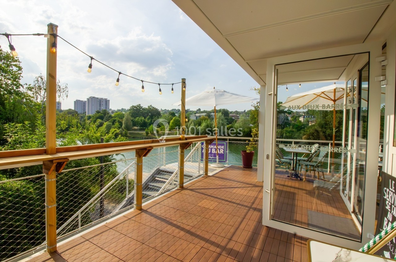 Terrasse en bois avec guirlandes lumineuses, vue sur une rivière bordée de végétation et des immeubles au loin.