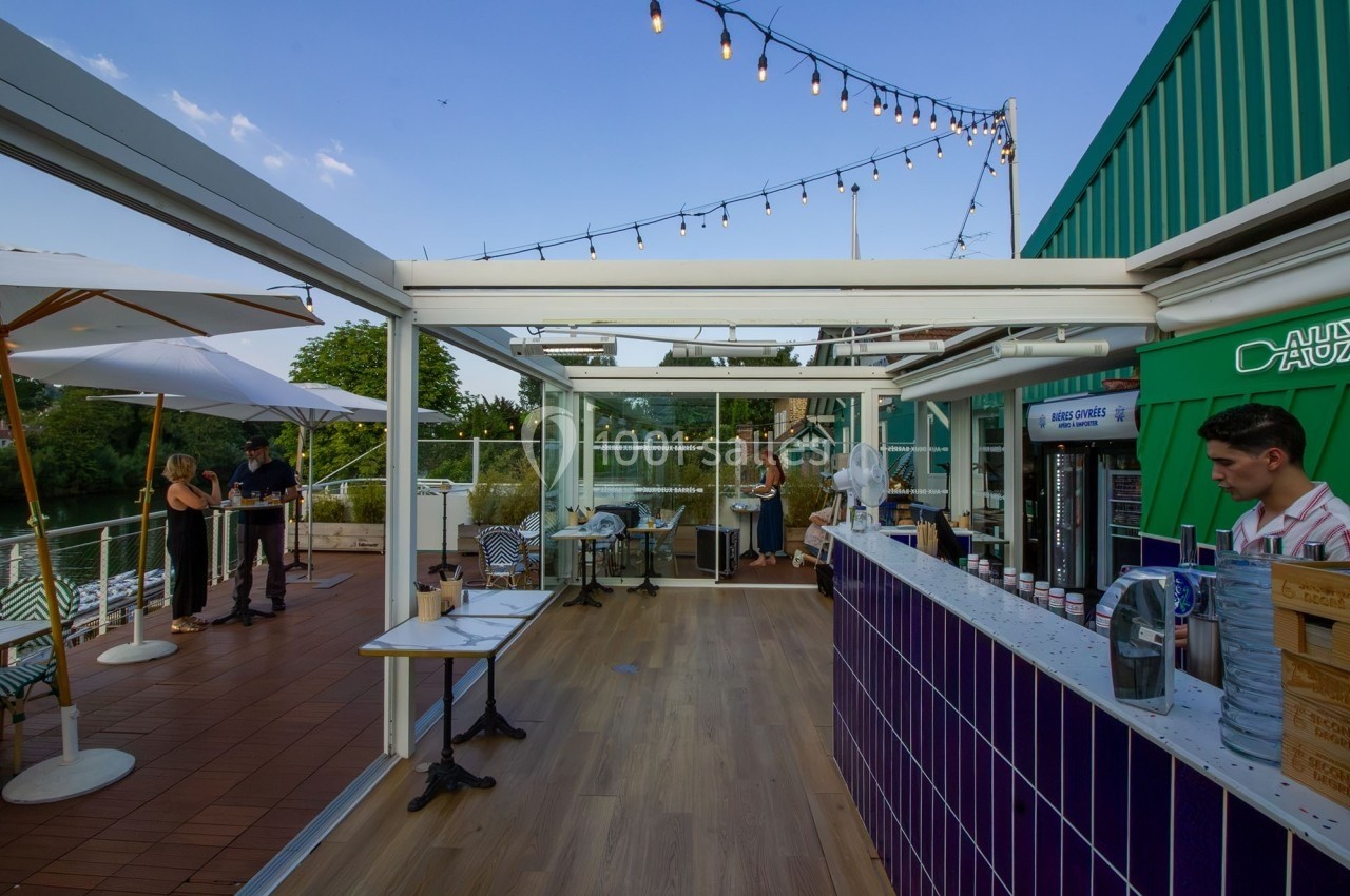 Terrasse extérieure avec bar en carrelage violet, tables, parasols et guirlandes lumineuses sous un ciel dégagé.