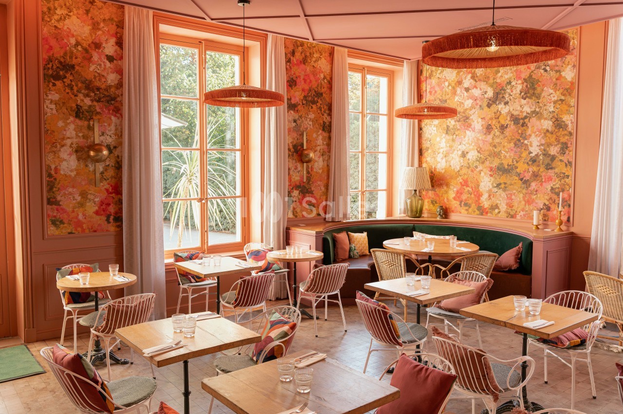 Salle de restaurant aux murs fleuris orange et rose, tables basses en bois, chaises cannées, baies vitrées sur jardin.