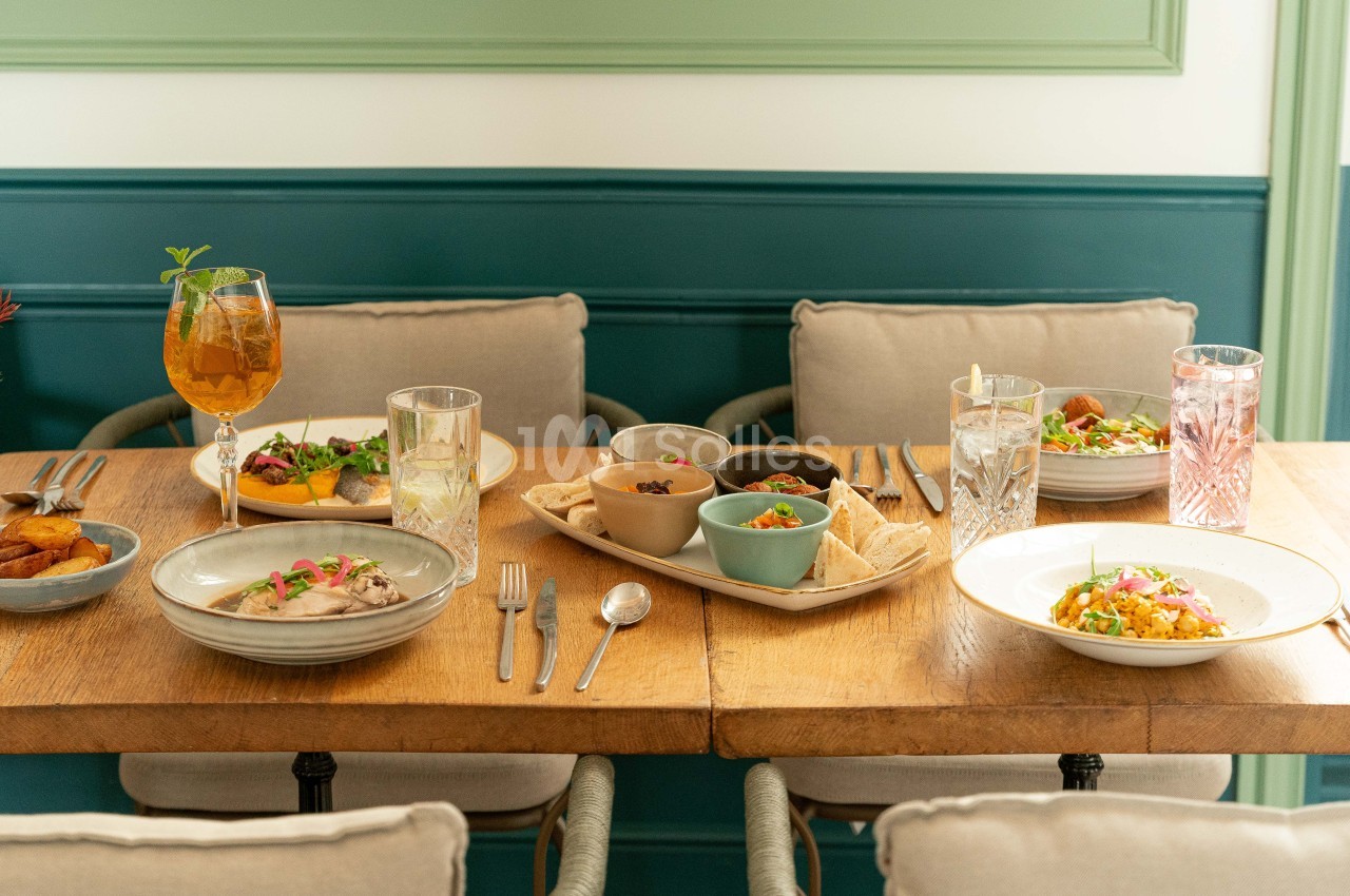 Table dressée en restaurant avec plats cuisinés, verres et couverts face à banquette vert foncé.