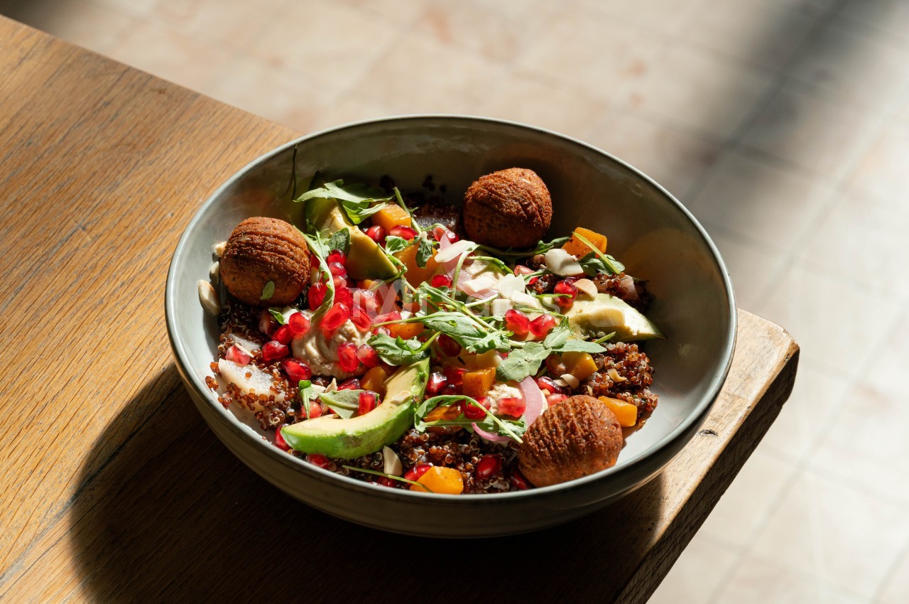 Bol en céramique contenant quinoa, boulettes panées, légumes colorés et herbes fraîches sur plateau en bois.