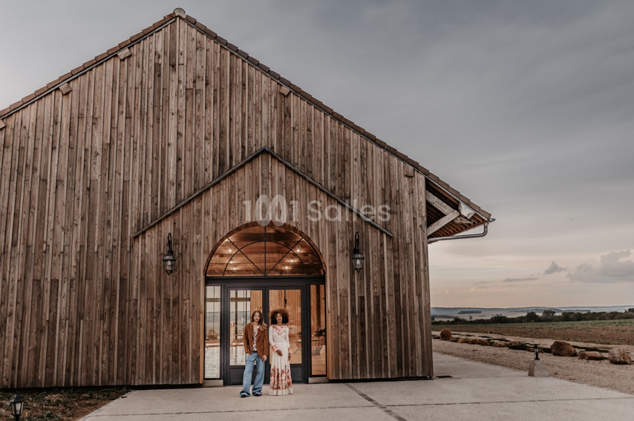 Bâtiment en bois avec entrée cintrée et éclairage intérieur - couple devant la façade en fin d'après-midi.