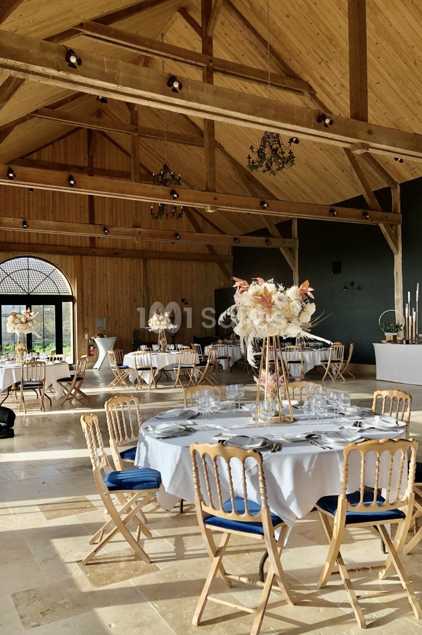 Salle aux poutres apparentes avec tables rondes dressées, chaises en bois naturel à coussins bleus et compositions florales.