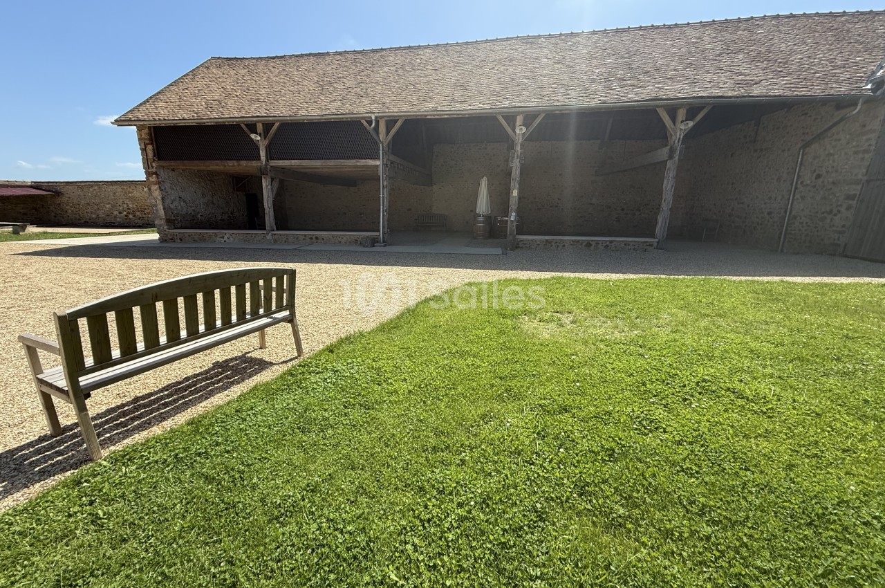 Cour de ferme avec hangar ouvert aux poutres blanches, pelouse verte et banc en bois.