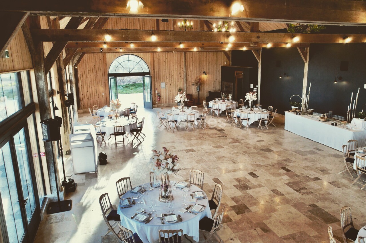 Salle en grange aménagée avec poutres apparentes, tables rondes dressées, chaises chiavari et éclairage guirlande - ouverture sur parc.