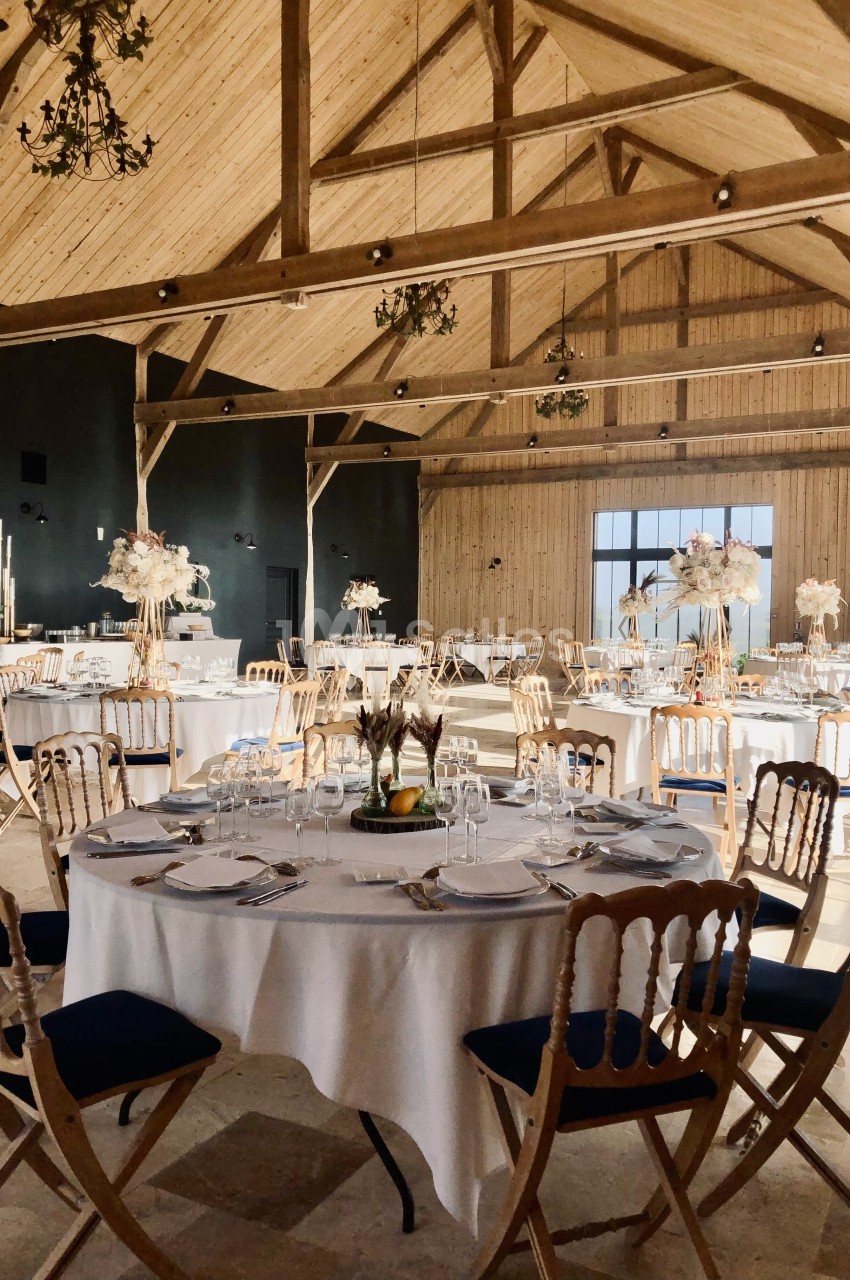 Salle de réception rustique dressée avec tables rondes blanches, chaises en bois et fleurs blanches.