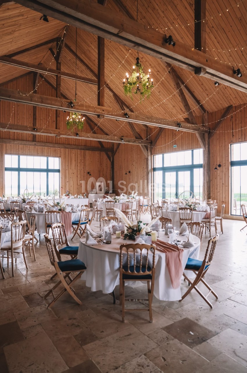 Grange en bois aménagée pour réception - tables rondes dressées, chaises en bois, lustres floraux, guirlandes lumineuses.