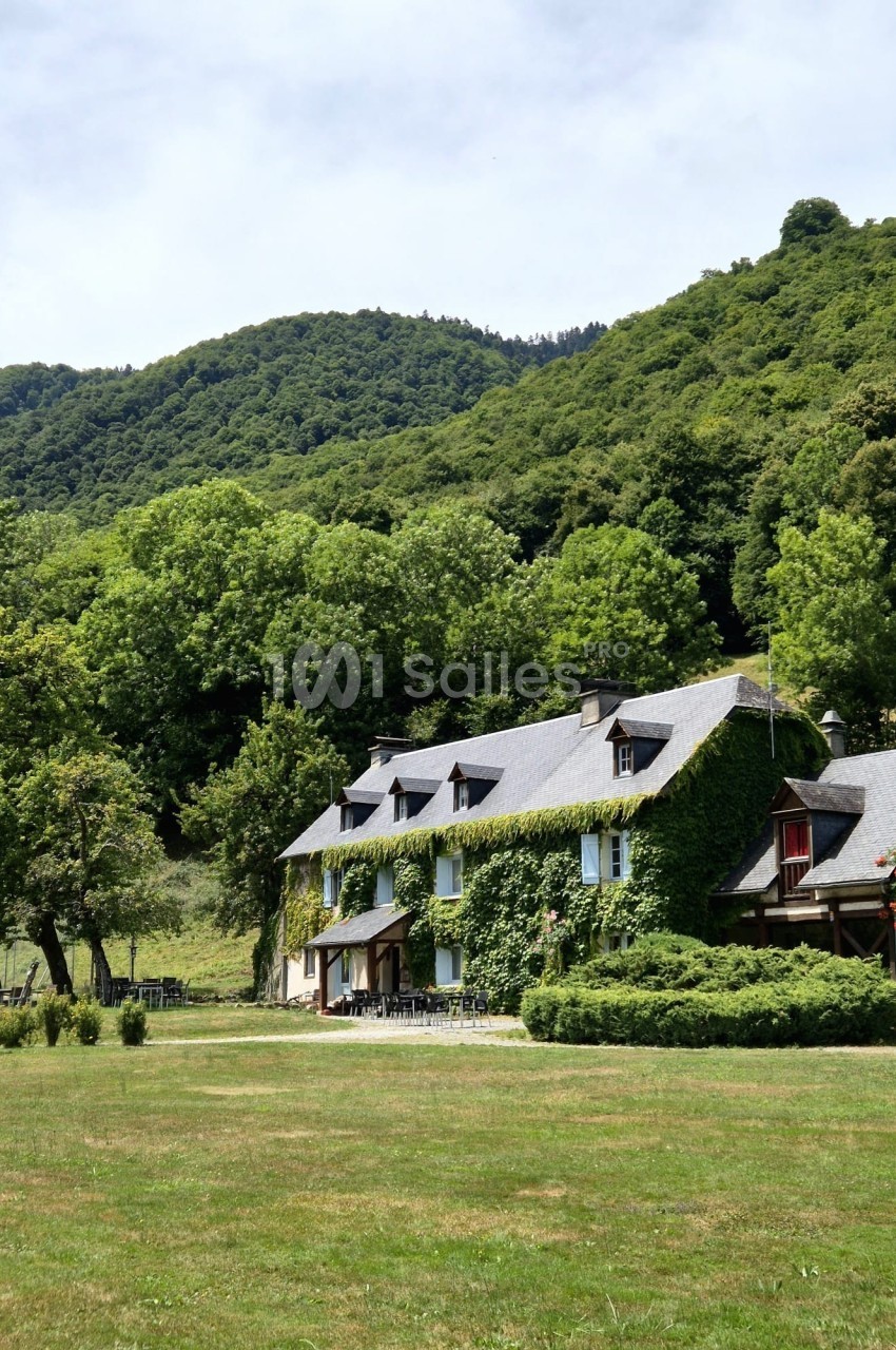 Bâtiment en pierre couvert de lierre avec toit en ardoise et lucarnes, au pied d'une colline boisée, devant une grande pelouse et des tables dressées en terrasse.