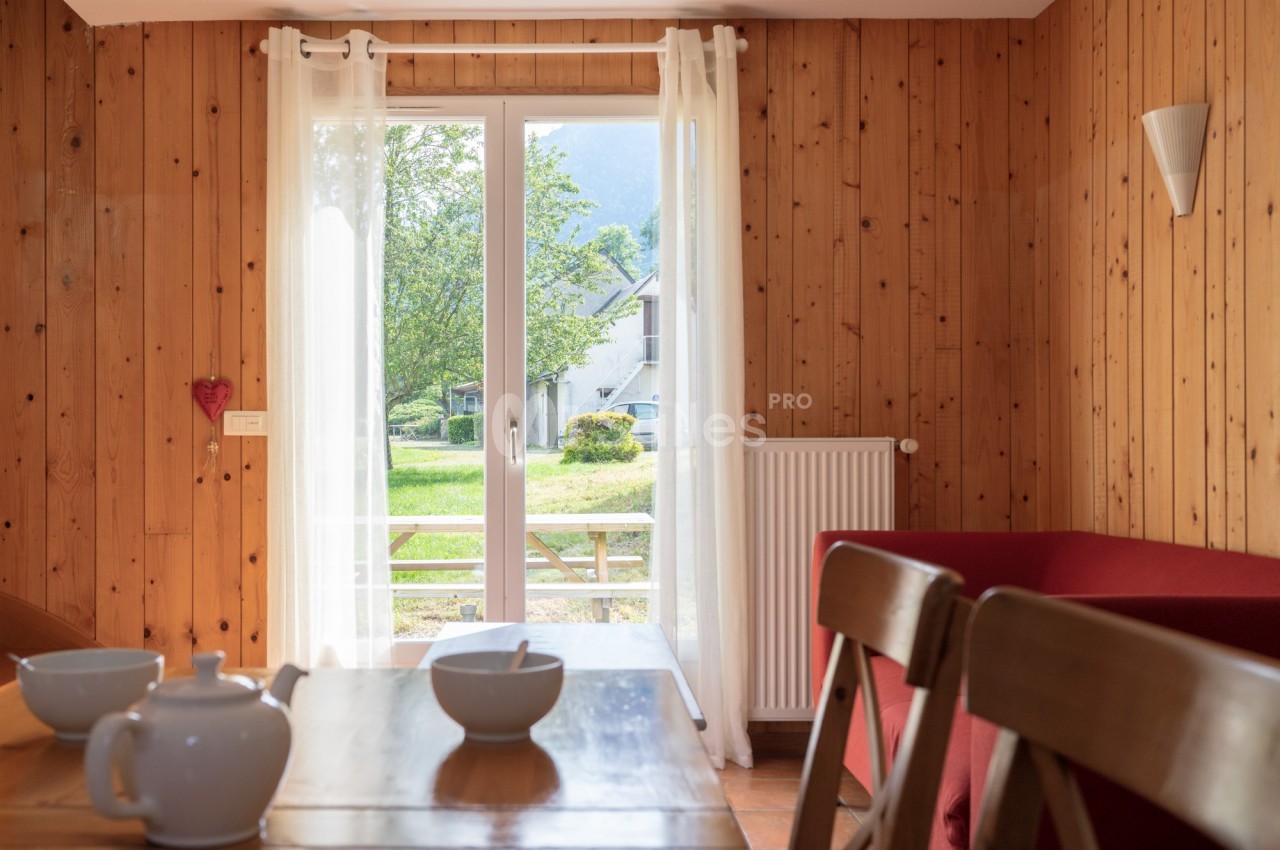 Pièce aux murs et plafond en lambris de bois clair, avec une table en bois dressée de bols et d'une théière blanche, une banquette rouge et une porte-fenêtre donnant sur un jardin verdoyant.