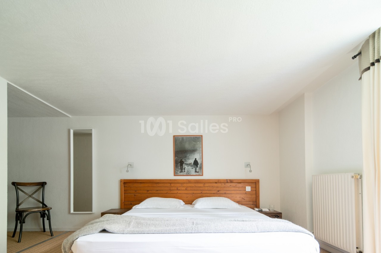 Chambre aux murs blancs avec un grand lit à tête de lit en bois clair, deux chevets, une chaise en bois et métal et un radiateur blanc.