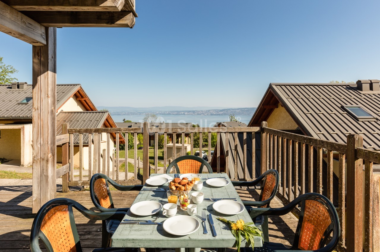Terrasse en bois dressée pour quatre personnes avec viennoiseries, jus et tasses, donnant sur des maisons mitoyennes et une vue dégagée sur le lac.