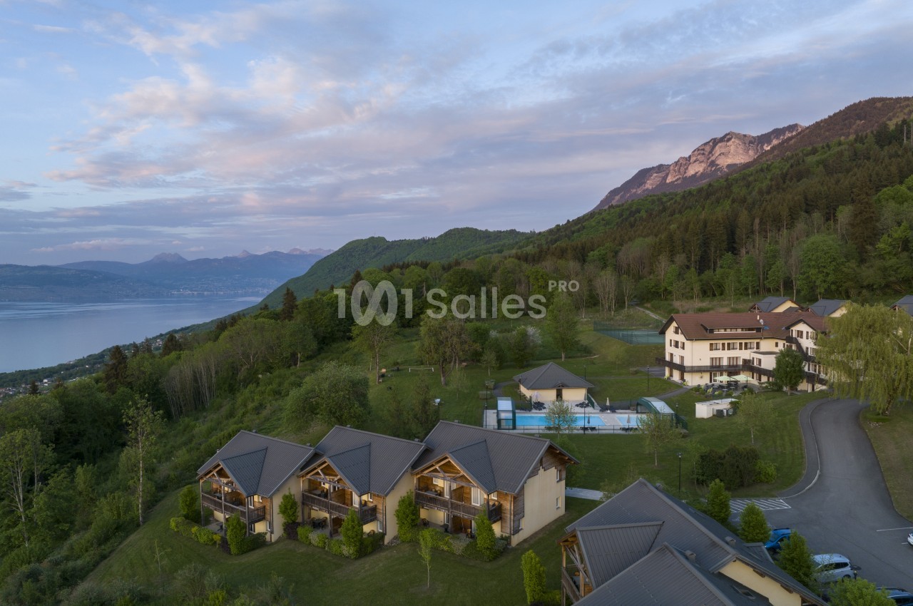 Vue aérienne d'un ensemble de chalets en bois et d'un bâtiment principal blanc au bord d'une pente boisée, avec piscine et lac en arrière-plan.