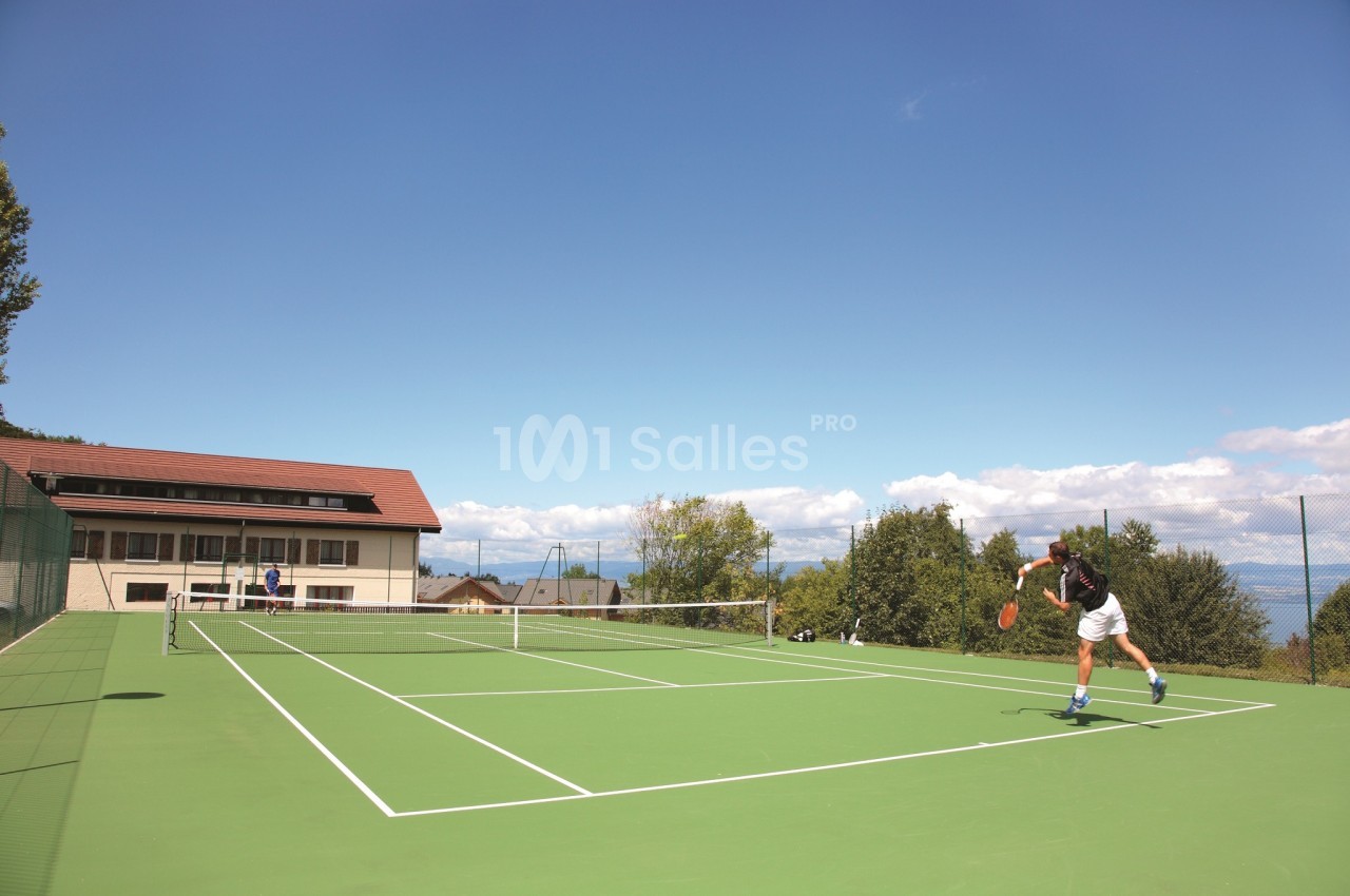 Court de tennis en extérieur à gazon synthétique vert, avec un joueur en plein service et un bâtiment d'un étage à toit de tuiles sur la gauche.