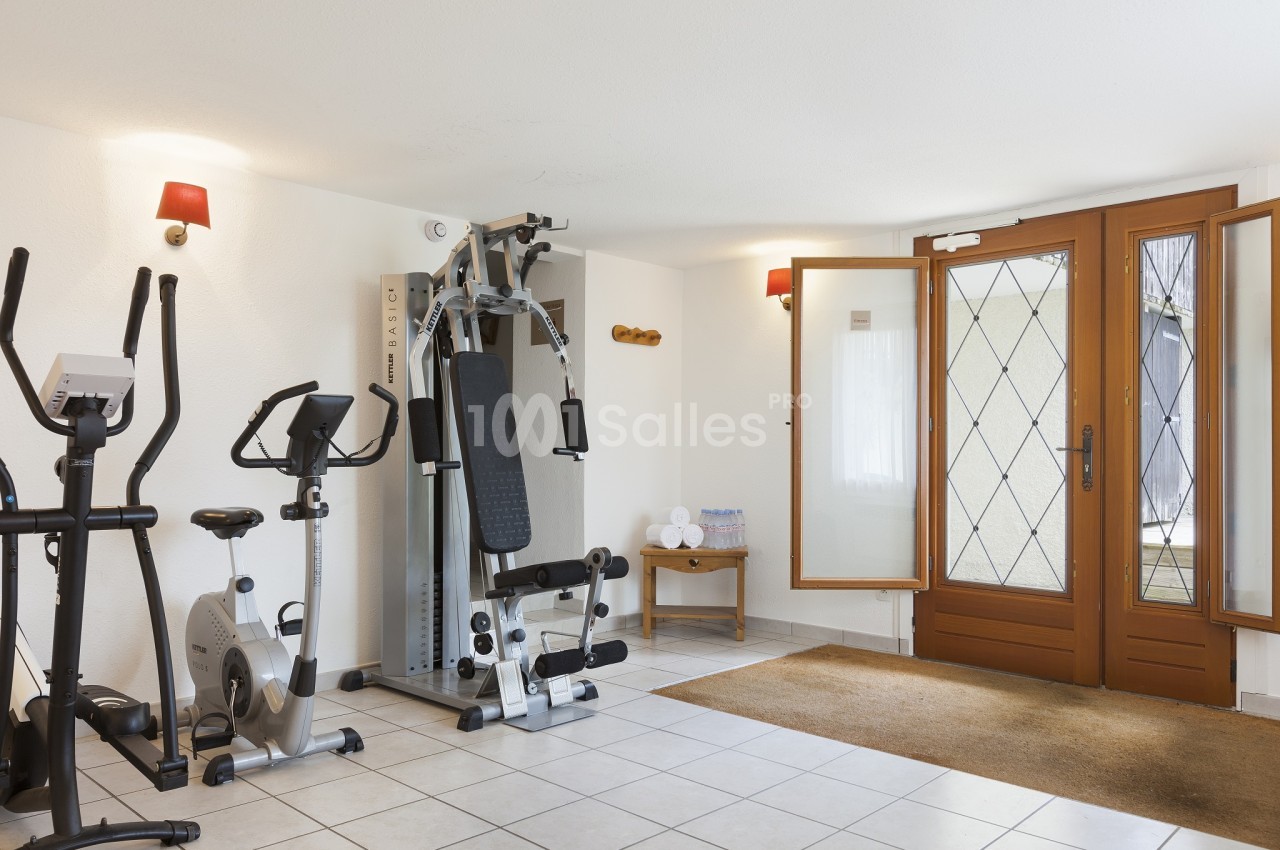 Salle de sport aménagée avec un vélo elliptique, un vélo d'appartement et un appareil de musculation, carrelage clair et portes-fenêtres en bois vitrées.