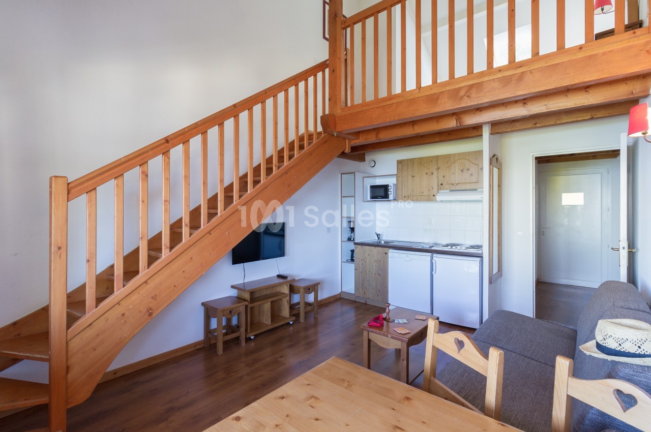 Séjour en bois clair avec escalier menant à une mezzanine, coin cuisine équipé à droite, canapé gris et table en bois au premier plan.