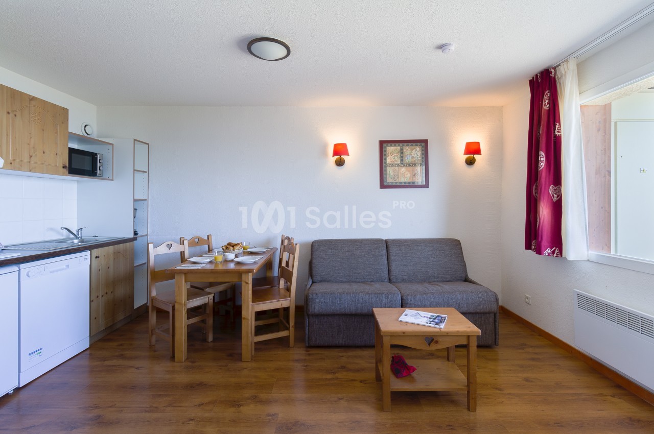 Séjour d'un appartement avec coin repas en bois à quatre chaises, canapé gris, cuisine équipée sur la gauche et rideaux rouges à motifs. Let me check length: roughly 150 characters, too long. Need ≤125. Séjour meublé avec table en bois et chaises, canapé