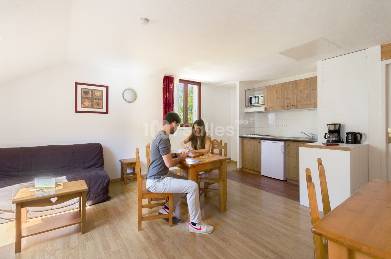 Séjour lumineux d'un appartement avec coin salon à gauche, table en bois où deux personnes sont assises à jouer, et cuisine équipée à droite.