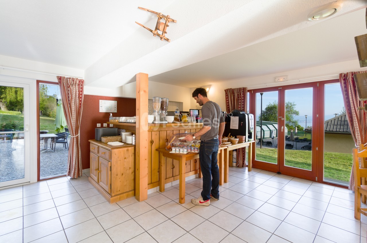 Intérieur lumineux avec carrelage clair : un homme se sert au buffet du petit-déjeuner, sur un meuble en bois, près de grandes portes-fenêtres donnant sur une pelouse.
