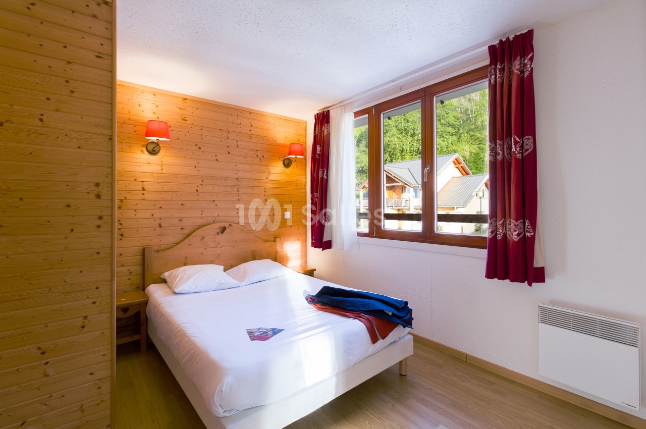 Chambre aux murs et plafond en bois clair, avec un grand lit double à tête en bois, deux appliquets rouges et des rideaux rouges à motifs. Let me check length: roughly 135 characters? Count: "Chambre aux murs et plafond en bois clair, avec un grand lit