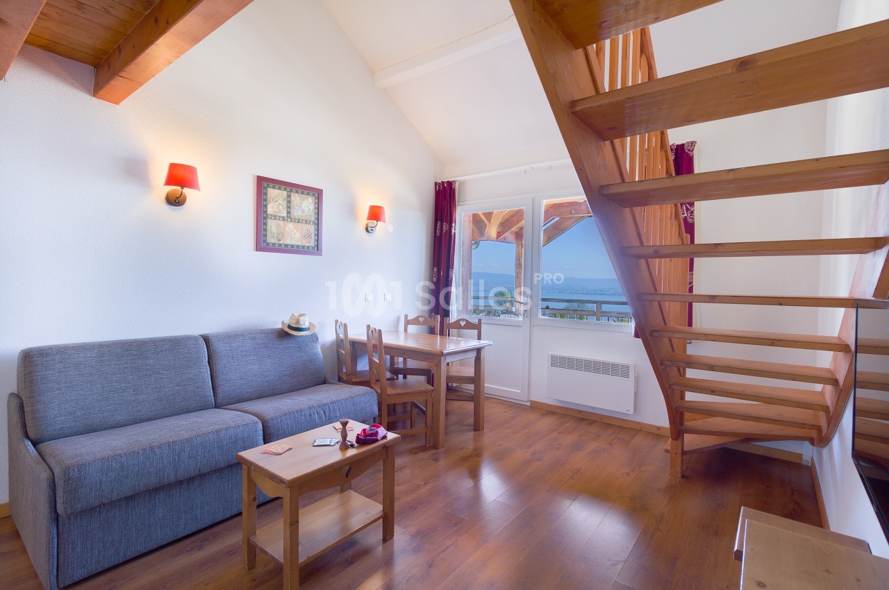 Séjour en bois clair avec canapé gris, table et chaises en bois, escalier menant à l'étage et grandes fenêtres donnant sur un paysage lacustre.