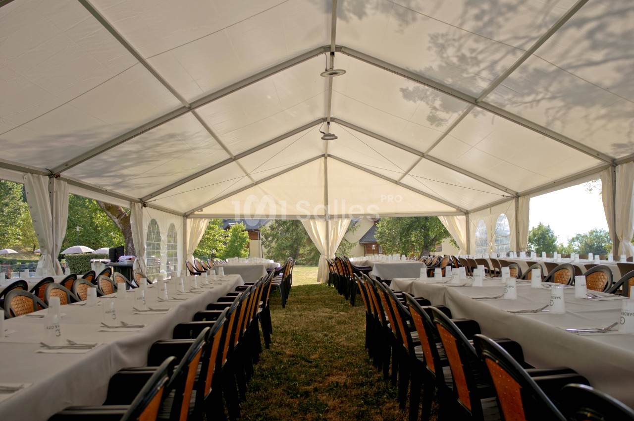 Grande tente blanche sous laquelle sont dressées de longues tables nappées de blanc avec chaises en bois et métal, sur une pelouse, en extérieur.