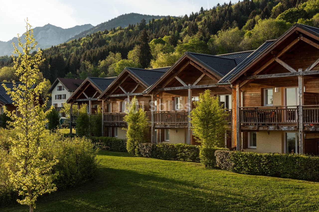 Bâtiments en bois à toits à deux pans sombres, avec balcons et pelouse, au pied d'une forêt et de montagnes en arrière-plan, en fin de journée.